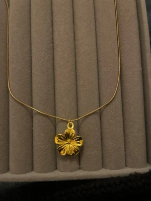 Guldigt halsband med blomma - Snyggt halsband i guldfärg med en blomformad berlock. Kedjan är tunn och elegant med justerbar längd och karbinlås. Blomman har fina detaljerade blad och ger ett stilrent intryck. Perfekt accessoar för att lyfta din outfit.