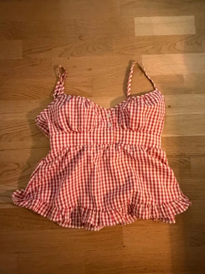 Rutig röd och vit linnetopp - Supersöt linnetopp med rödvitt rutigt mönster och smala axelband. Toppen har volangkant nedtill och smockad rygg för extra bekväm passform. Perfekt för varma dagar och ger en gullig vintagekänsla.