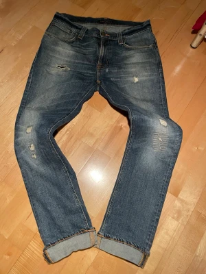 Nudie jeans - Thin finn 32/33  - Säljer ett par blå jeans från Nudie Jeans med slitningar och distressed detaljer på benen. Jeansen har klassisk femficksmodell, orangea sömmar och uppvikta benslut. Tillverkade i denim med snygg tvätt och vintagekänsla. Sjukt snygga och trendiga med galet snygg tvätt och slitningar. 32/33. Sjukt snygga. Skriv vid funderingar 