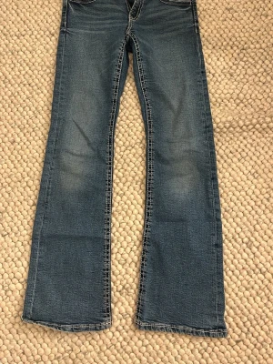 Blå bootcut jeans från Gina Tricot Young - Snygga blå bootcut jeans från Gina Tricot med vita kontrastsömmar och dubbla knappar i midjan. Jeansen har klassisk femficksdesign och lätt slitning framtill för en cool look. Perfekta för dig som gillar en avslappnad och trendig stil. i bra skick inte använd särskilt mycket.