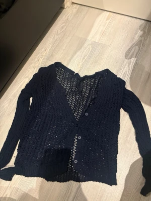 Mörkblå virkad kofta med knappar - Snygg mörkblå kofta med virkat mönster och v-ringning. Koftan har långa ärmar och stängs med fyra knappar framtill. Perfekt att slänga över en topp för en chill look. Materialet känns lätt och luftigt.