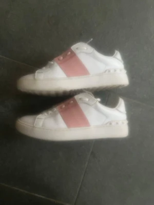 Valentino vita sneakers med rosa band - Snygga vita sneakers från Valentino med ett brett rosa band över mitten. Skorna har klassisk låg profil, rund tå och vita snören. Tillverkade i läder med gummisula och subtila nitar bak för extra edge. Väldigt bra skick, använts få gånger 🥰🥰