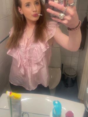 Rosa volangblus med v-ringning - Söt ljusrosa blus med volanger och djup v-ringning. Blusen har korta ärmar och är gjord i ett glansigt, lätt material som ger en romantisk känsla. Perfekt för dig som gillar feminina detaljer och en trendig look.