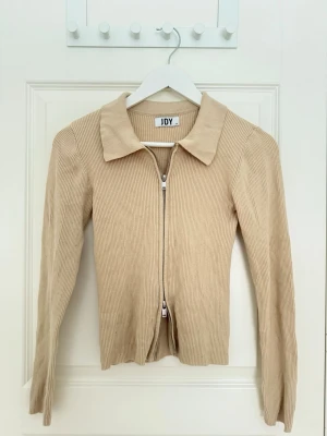 Beige zip cardigan med krage - Beige zip kofta som går att dra åt båda hållen. Koftan är ribbad med justerbar dragkedja och en krage. 