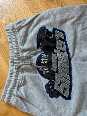 Gråa mjukisbyxor från Trapstar - Säljer ett par gråa mjukisbyxor från Trapstar med stor broderad logga och texten 'Shooters' på ena benet. Byxorna har dragsko i midjan och resår vid benslut. Perfekta för en chill och street-inspirerad look.