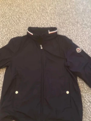 Svart Moncler jacka - Snygg svart jacka från Moncler med dragkedja framtill och två fickor med tryckknappar. Jackan har hög krage med rödvit detalj och Moncler-logga på ärmen. 