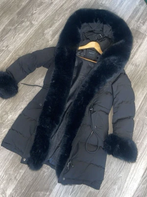 Svart pufferkappa  - Snygg svart pufferkappa från Copperose med stor huva och breda detaljer i fuskpäls längs huva och ärmslut. Kappan har knytband i midjan och praktiska fickor med lock. Perfekt för kalla dagar när du vill hålla stilen och värmen. Köpt för 1800kr 