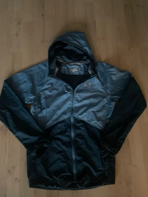 Mörkblå  och blå vindjacka Under Armour - Snygg mörkblå  och blå  vindjacka från Under Armour med huva och dragkedja framtill. Jackan har en sportig design, meshfoder och är tillverkad i lätt, vindtätt material. Perfekt för löpning eller utomhusaktiviteter. Under Armour-logga på bröstet.