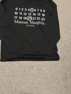 Maison Margiela svart långärmad tröja - Svart långärmad tröja från Maison Margiela med ikoniskt siffertryck i vitt framtill och rund halsringning. Tröjan har en avslappnad passform och är tillverkad i mjukt bomullsmaterial. Perfekt för dig som gillar streetwear och unika designer. Kan passa M me!