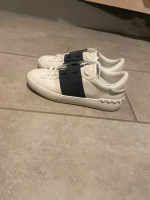Vita sneakers med marinblå detalj - Snygga vita sneakers med en bred marinblå remsa över mitten och matchande blå snörhål. Skorna har en låg profil och är tillverkade i slätt läder med en stilren design. Perfekta för dig som gillar minimalistisk och modern stil.