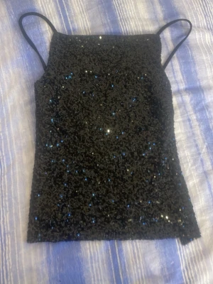 Svart paljettlinnetopp med smala band - Snygg svart linnetopp täckt med glittriga paljetter och smala axelband. Toppen har justerbara band på baksidan för perfekt passform och en croppad modell som passar till många olika looks. Perfekt för dig som vill sticka ut lite extra.