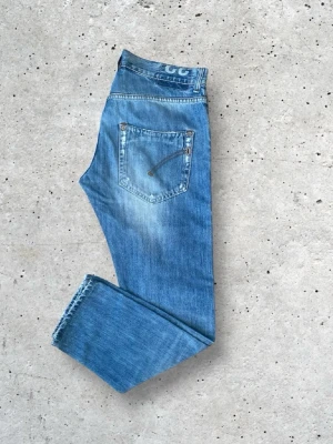 Blå raka jeans från dondup med slitningar - Säljer ett par dondup blå jeans med rak passform och snygga slitningar på framsidan. Jeansen har klassisk femficksdesign och detaljerade sömmar på bakfickan. Tillverkade i denim med en cool tvättad look, perfekta för en avslappnad stil.