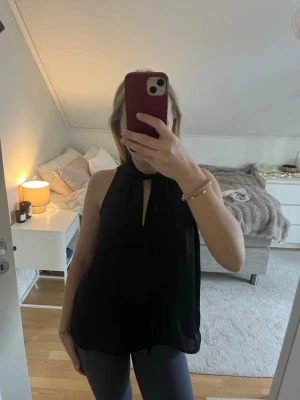 Svart ärmlös blus med knyt - Säljer en svart, ärmlös blus med knyt i halsen och en liten slits framtill. Blusen har en luftig och flowy passform. 