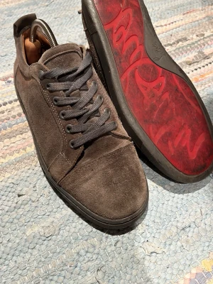 Christian louboutin gråa - Snygga sneakers i grå mocka med gråa skosnören och en röd sula med mönster. Klassisk låg modell med rund tå och diskret design som passar till det mesta. Skon har en liten defekt på den vänstra skon men går ej igenom till insidan av skon. Perfekta för dig som gillar stilrena detaljer och vill ha något lite annorlunda. Skulle behövas fräschas upp lite bara. 