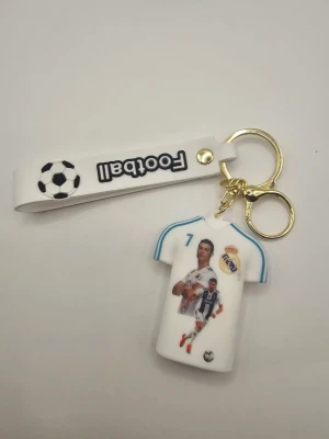 Nyckelring med Ronaldo-tröja - Cool nyckelring med vit Real Madrid-tröja, nummer 7 och texten 'RONALDO' på baksidan. Framsidan har bilder av fotbollsspelare och klubbens logga. Medföljer vit rem med fotbollsmotiv och texten 'Football'. Metallring i guld. Perfekt för fotbollsfans.