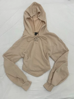 Beige cropped hoodie med snörning - Snygg beige cropped hoodie från Bershka med huva och snörning framtill. Hoodien har långa ärmar med breda muddar och är gjord i mjukt sweatshirtmaterial. Perfekt för en avslappnad och trendig look.