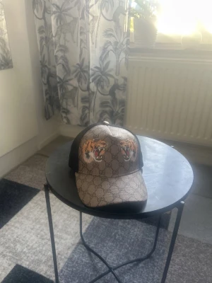 Gucci keps med tigertryck - Snygg keps från Gucci med klassiskt GG-mönster i beige och brunt samt två färgstarka tigerhuvuden framtill. Kepsen har svart mesh baktill och justerbar rem. Materialet är en mix av canvas och mesh, perfekt för en trendig streetstyle-look.