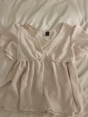 Ljusrosa blus med puffärm och knappar - Söt ljusrosa blus med v-ringning och knappar framtill. Blusen har korta puffärmar och är dekorerad med små broderade prickar. Köpt på Plick men originellt från Shein🤗!!Om du köper välj gärna att hämta vid upphämtningsställe istället för hemleverans då det blir enklare för mig att posta på det viset!!