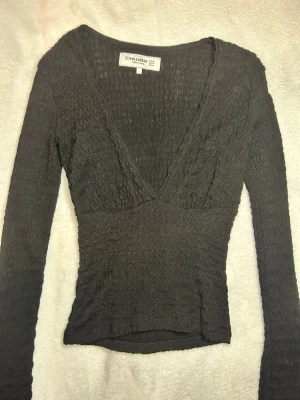 Svart v-ringad topp från Pull&Bear - Säljer en svart långärmad topp från Pull&Bear med v-ringning och smockad struktur. Toppen har en figurnära passform och är perfekt att styla med jeans eller kjol. Enkel men trendig design som passar till många tillfällen.