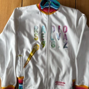 Retro ”Adidasjacka” i färgsprakande design - Säljer en unik vit jacka från Misericordia med färgglada bokstäver och mikrofontryck. Jackan har en dragkedja framtill och regnbågsfärgade muddar vid krage, ärmslut och nederkant. Perfekt för dig som vill sticka ut med stil! 🎤