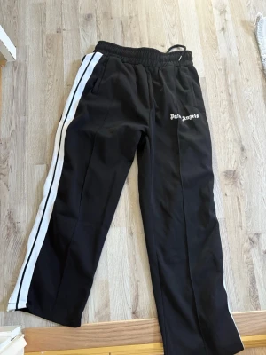 Palm Angels svarta mjukisbyxor - Palm Angels svarta mjukisbyxor med vita sidoränder och logga på vänster lår. Byxorna har resår i midjan med dragsko och raka ben. Perfekta för en avslappnad och trendig streetwear-look.