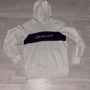 Vit hoodie från GANT med logga - Snygg vit hoodie från GANT med stor svart och röd rand över bröstet och tydlig GANT-logga. Klassisk känguruficka framtill och huva. Tillverkad i mjukt material som känns skönt mot huden. Perfekt för en avslappnad och trendig look.
