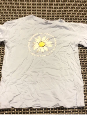 Ljusblå t-shirt med prästkrage - Ljusblå t-shirt med tryck av en stor prästkrage i vitt och gult på bröstet. Runt blomman står texten 'All flowers of tomorrow are in the seeds of today'. Klassisk passform och rund hals. Perfekt för dig som gillar blommiga motiv.