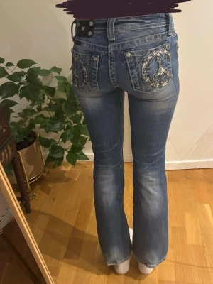 Miss me Blå bootcut jeans  - Säljer ett par blå miss me bootcut jeans med slitningar och peace-märken i paljetter på bakfickorna. Jeansen har låg midja, femficksmodell och detaljer vid fickorna. Midjemått: 33cm.🩷 Innerbenslängd: 74cm.🩷
