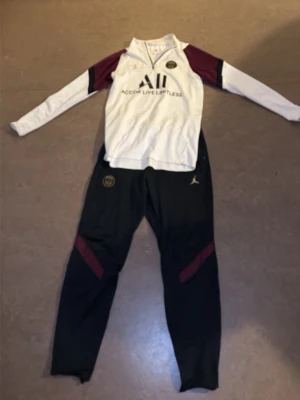 PSG x Jordan träningsset svart/vit - Snyggt PSG x Jordan träningsset med långärmad vit tröja med vinröda detaljer och svart byxa med vinröda inslag. Tröjan har dragkedja vid halsen och tryckta loggor. Byxorna har elastisk midja, PSG-märke och Jumpman-logga. Perfekt för fotbollsträning eller chill.