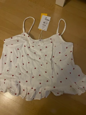 Vit pyjamaslinne med röda hjärtan - Sött vitt pyjamaslinne med tunna axelband och små röda hjärtan över hela tyget. Linnet har volangkant nedtill och är gjort i ett lätt och luftigt material, perfekt för mysiga kvällar hemma. Aldrig använt