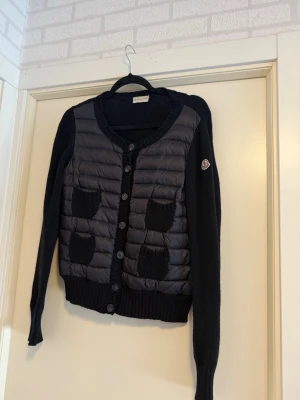 Svart cardiganjacka från Moncler - Snygg svart cardiganjacka från Moncler med quiltad front och stickade ärmar. Fyra små fickor framtill och Moncler-logga på ärmen. Jackan har rund halsringning och knäppning med knappar. Storlek M men sitter lite mindre, passar bäst på S