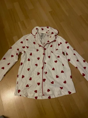 Vit pyjamaströja med röda hjärtan - Supersöt vit pyjamaströja med röda hjärtan över hela och röda kanter längs knäppningen. Skön och mjuk, perfekt för mysiga kvällar hemma. Långärmad och med klassisk krage. Materialet är bomull. Tröjan har en sminkfläck vid kragen men går säkert bort i tvätten. Använd en gång.