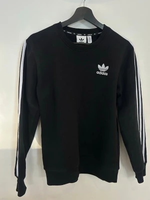 Svart Adidas sweatshirt med ränder - Klassisk svart sweatshirt från Adidas med vita ränder längs ärmarna och Adidas-logga på bröstet. Tröjan har rund halsringning, långa ärmar och ribbade muddar. Perfekt för en sportig och avslappnad stil. Storlek M man, jag som tjej 160cm använder den som over sized myströja💞