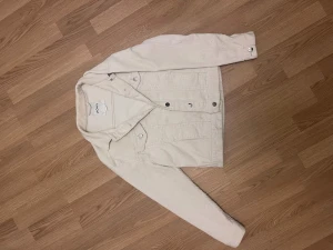 Vit jeansjacka från MONKI - Snygg vit jeansjacka från MONKI med klassisk krage, silverfärgade knappar och två bröstfickor med lock. Jackan har raka ärmar och en avslappnad passform.