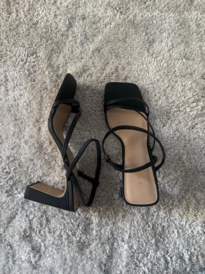 Svarta pumps med remmar från H&M - Stilrena svarta pumps från H&M med fyrkantig tå och tunna remmar över foten. Skorna har en bred klack och öppen häl, vilket ger en modern look. Tillverkade i konstläder med beige innersula för extra kontrast.