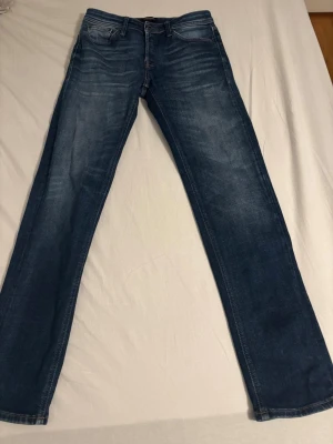Blå raka jeans med slitningar - Säljer ett par klassiska blå jeans med raka ben och diskreta slitningar framtill. Jeansen har fem fickor och normal midja. Materialet är denim i bomull och de har en snygg tvättad look. Perfekta till en avslappnad stil.