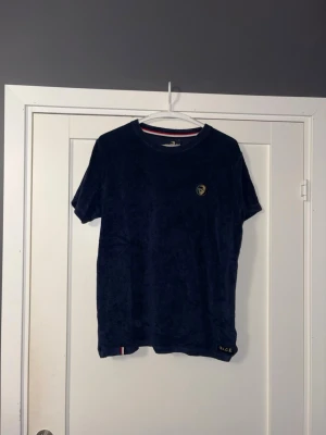 Mörkblå frotté t-shirt från Pace - Mörkblå t-shirt från Pace i mjuk frotté med gul broderad logga på bröstet. Klassisk rund halsringning och korta ärmar. Liten Pace-detalj vid nederkanten. Perfekt för dig som gillar unika material och stilrena detaljer.