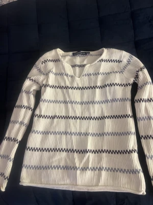 Vit stickad tröja med blå ränder - Säljer en vit ribbstickad tröja från Alan Paine med tunna horisontella ränder i blått och grått. Tröjan har v-ringning och långa ärmar, perfekt för en avslappnad och stilren look. Materialet känns mjukt och följsamt.