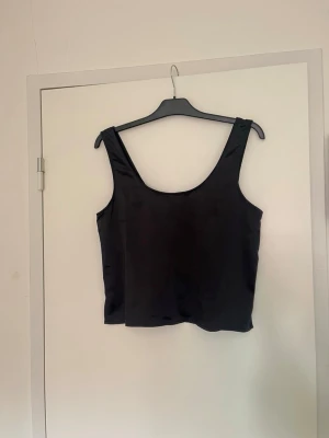 Svart linnetopp med breda axelband - Snygg svart linnetopp med breda axelband och rundad urringning. Toppen har en enkel och stilren design som passar till många olika looks. Materialet känns mjukt och har en lätt glansig finish.