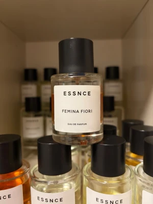 ESSNCE Femina Fiori Eau de Parfum - ESSNCE Femina Fiori Eau de Parfum kommer i en rund, genomskinlig glasflaska med svart plastlock och enkel vit etikett. Doften har en lätt rosa ton som syns genom flaskan. Minimalistisk och stilren design som känns modern och fräsch.