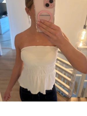 Vit bandeau topp med volangkant - Säljer en vit bandeau topp från bikbok i storlek xs med smockad midja och volangkant nedtill. Toppen är axelbandslös och har en mjuk, stretchig passform som sitter snyggt på kroppen. Perfekt till jeans eller kjol för en fräsch look💞