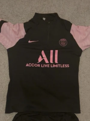 PSG träningströja och byxor - M passar som S. Snygg svart PSG träningströja och byxor från Nike med rosa detaljer och logga. Tröjan har halv dragkedja, rosa ärmar och Paris Saint-Germain klubbmärke på bröstet. Tillverkad i funktionsmaterial som passar perfekt till träning.