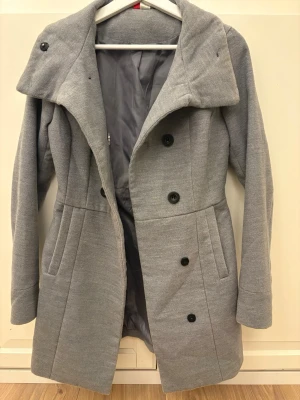 Ljusgrå dubbelknäppt kappa - Snygg ljusgrå kappa med dubbelknäppning och bred krage. Kappan har två fickor framtill och är helfodrad för extra komfort. Stängs med svarta knappar och har en modern, rak passform som passar till många olika stilar. Kappan är lite längre och hyfsat liten i storleken