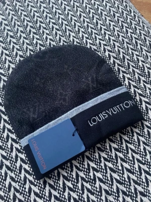 Svart mössa från Iv - Snygg svart mössa från Iv med diskret monogrammönster och en ljusgrå rand vid kanten. Märkets logga är broderad i vitt framtill. Tillverkad i mjukt stickat material som känns lyxigt och stilrent.