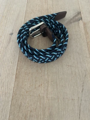 Blått flätat bälte med silverdetaljer - Snyggt flätat bälte i marinblått och ljusblått med silverfärgat spänne och bruna skinn-detaljer. Perfekt accessoar för att lyfta din outfit med en cool touch. Bältet har en flexibel design som gör det enkelt att justera.