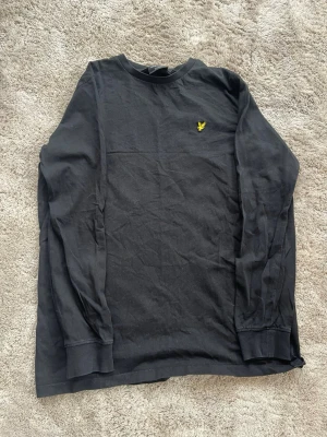 Svart långärmad tröja Lyle & Scott - Svart långärmad tröja från Lyle & Scott med klassisk gul logga broderad på bröstet. Tröjan har rund halsringning och ribbade muddar vid ärmslut och nederkant. Tillverkad i mjukt bomullsmaterial, perfekt för en avslappnad stil. OBS! Storlek passar 15-16 år.