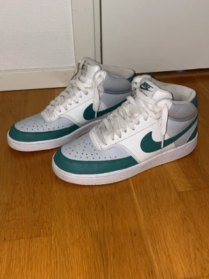 Nike vita och gröna midsneakers - Nike sneakers i vitt och grönt med klassisk swoosh på sidan. Modellen är mid och har perforerad tåbox, vita skosnören och vadderad plös. Tillverkade i läder och syntetmaterial. Snygg retrostil som passar till många outfits.
