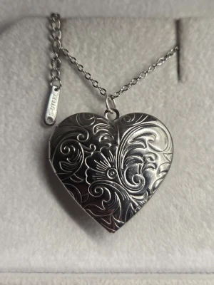 Silverfärgat medaljong hjärthalsband med mönster - Unikt halsband i silverfärg med ett hjärtformat medaljonghänge. Medaljongen har vackra, detaljerade blommönster och kan öppnas för att lägga in ett foto eller minne. Kedjan är tunn och är i rostfritt stål 45 cm. Perfekt accessoar för en personlig touch.