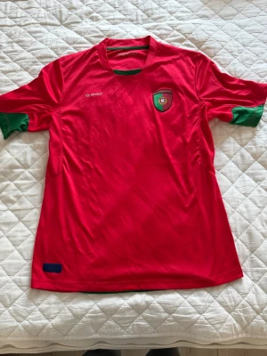 Portugal supportertröja röd-grön - Säljer en röd Portugal supportertröja från Soc med gröna detaljer vid ärmar och hals. Tröjan har Portugal-emblemet på bröstet och en blå patch med texten 'Official Supporter' nedtill. Tillverkad i lätt och ventilerande syntetmaterial. Den är använd ganska många gånger men ändå ganska bra skick