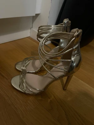 Metallic guldiga höga pumps - Säljer ett par metallic guldiga pumps med höga smala klackar och flera tunna remmar som går över foten och runt ankeln. Skorna har dragkedja bak för enkel påtagning och en öppen tå. Perfekta för dig som vill sticka ut med en glammig look.